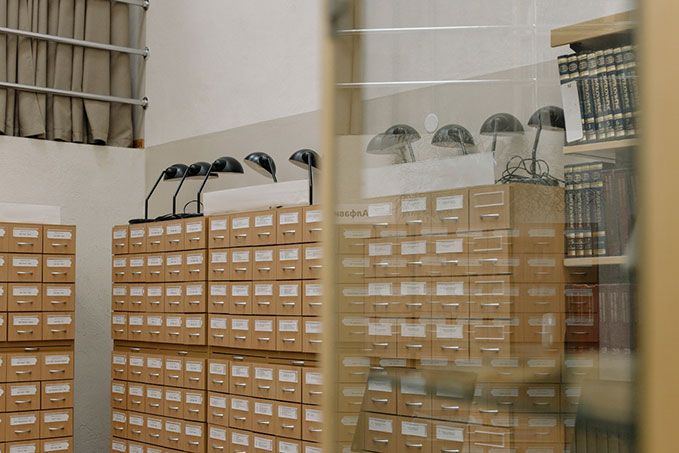 A corner of a white room. It’s full of tall, wooden filing cabinets with black lamps on top.