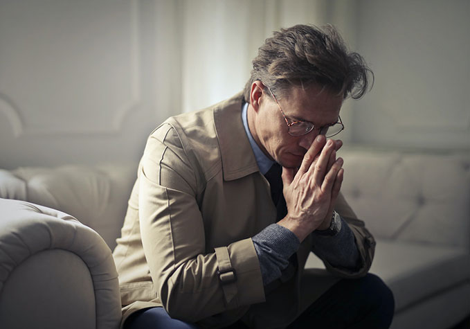 A middle-aged man in a tan trench coat sitting on a couch, looking pensive. He has his hands clasped in front of his face