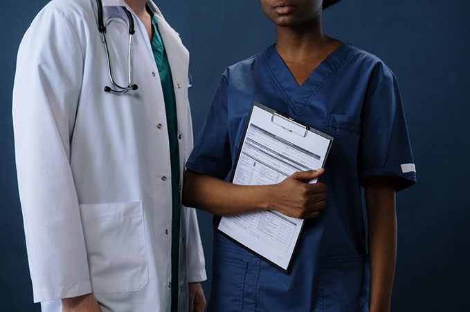 Two doctors’ side by side from the neck down. One is holding hospital paperwork, and the other has a stethoscope around their neck.