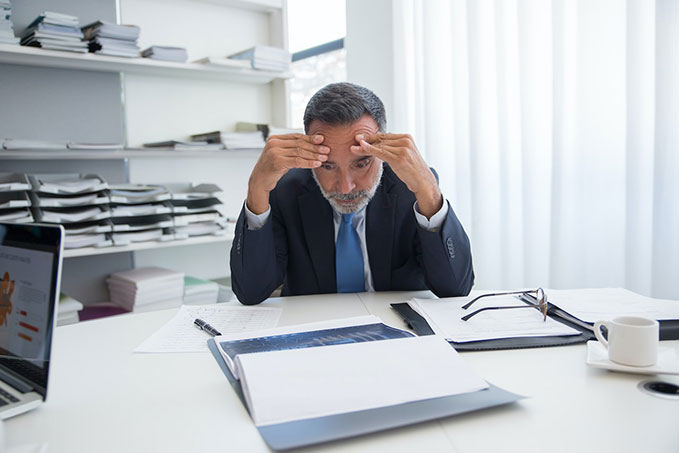 A businessman sat at a white desk surrounded by papers. He has his elbows on the desk and his hands are rubbing his temples. He looks concerned.