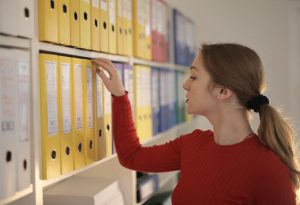 A lady wearing a red sweater browsing a shelf of many coloured folders and binders