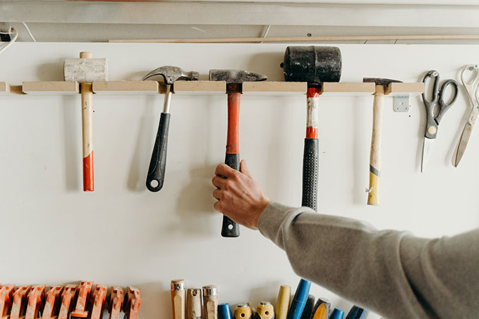 A row of five hammers hanging from a wooden shelf. An arm is reaching for the red, centre hammer