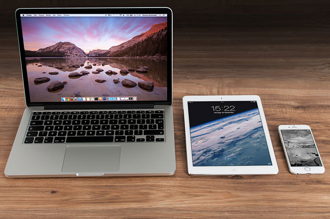 An open silver Mac laptop, a white tablet, and a white cellphone sit on a pine-coloured table.