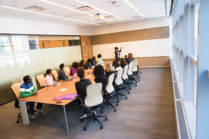 A view of a bright boardroom. There's a long desk in the middle with employees on either side. They're paying attention to a person writing on a whiteboard.