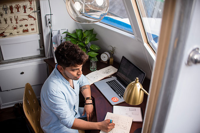 A photo looking down on a man in a blue shirt at a tidy desk with a laptop and a man sketching in a notepad